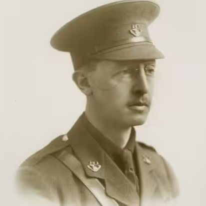 Sepia-toned portrait photograph of a man in World War I-era military uniform. He wears a peaked cap with insignia, a high-collared jacket with epaulettes, collar badges, and a cross-belt strap. He has a mustache and faces forward with a neutral expression against a plain light background.
