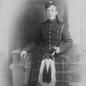 A black and white photograph of a young man dressed in a historical Scottish military uniform. He is wearing a kilt with a plaid pattern, a sporran hanging in front, and a double-breasted jacket with epaulets. He has a hat with a badge on his head and is holding a cane or swagger stick in his right hand. He is standing next to a stone pedestal, with one hand resting on it. 