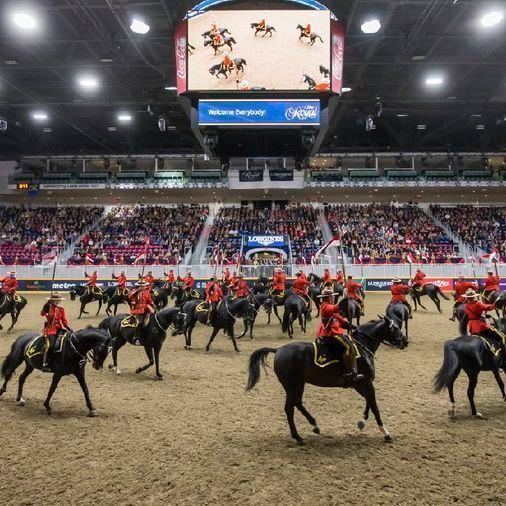 A large indoor arena filled with spectators in tiered seating. In the center ring, approximately 20 Royal Canadian Mounted Police officers in iconic red serge uniforms with yellow stripes and wide-brimmed Stetson hats perform the Musical Ride on black horses trotting in precise formation. Overhead, a big screen shows a close-up of riders and horses, with banners for Longines and other sponsors visible around the arena. Bright overhead lights illuminate the scene.