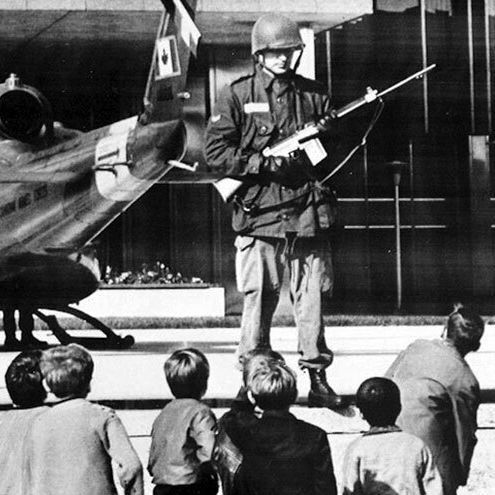 A black-and-white photo shows a soldier in uniform holding a rifle and standing in front of a helicopter, addressing a group of children seated on the ground. Another soldier is visible near the helicopter, which has a flag on its tail. The scene appears to take place in front of a modern building with large windows.