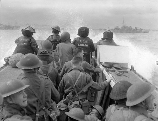 The black-and-white photo shows Canadian soldiers on a landing craft during the D-Day invasion of Normandy on June 6, 1944. The soldiers, wearing helmets and military gear, are preparing to disembark as the craft approaches the beach. Two crew members at the rear are marked with "R.C.N." (Royal Canadian Navy) and "L.C.O. 140" on their backs. Waves crash against the boat, and larger ships are visible in the background, indicating the scale of the operation. The atmosphere is tense, capturing a pivotal moment in World War II.