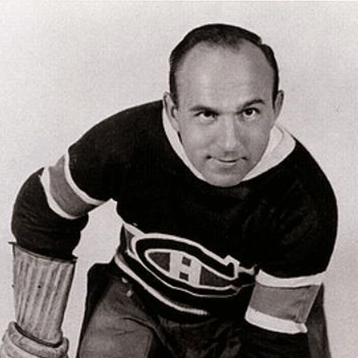 A black-and-white vintage photograph of Howie Morenz, a legendary Canadian ice hockey player, captured in a dynamic action pose during his career with the Montreal Canadiens. He is a middle-aged man with a bald head, intense eyes, and a determined smile, wearing a classic 1930s-era Canadiens jersey (black with white trim and the iconic red "CH" logo on the chest) and matching pants. His right arm is extended forward with a gloved hand gripping a hockey stick, while his left arm is bent at the elbow, as if skating aggressively toward the camera. The background is plain white, emphasizing his focused expression and athletic stance.