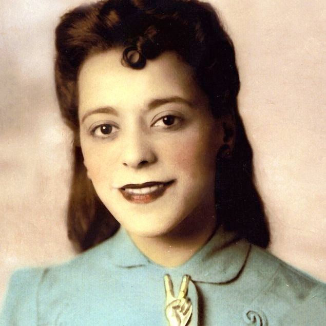 The photo depicts a person wearing a light blue garment with intricate embroidery on the shoulders. A prominent feature is a brooch shaped like a hand making a peace sign, positioned centrally on the chest. The background is a faded, neutral color, giving the image a vintage appearance. The person's hair is styled in soft curls, adding to the classic look.