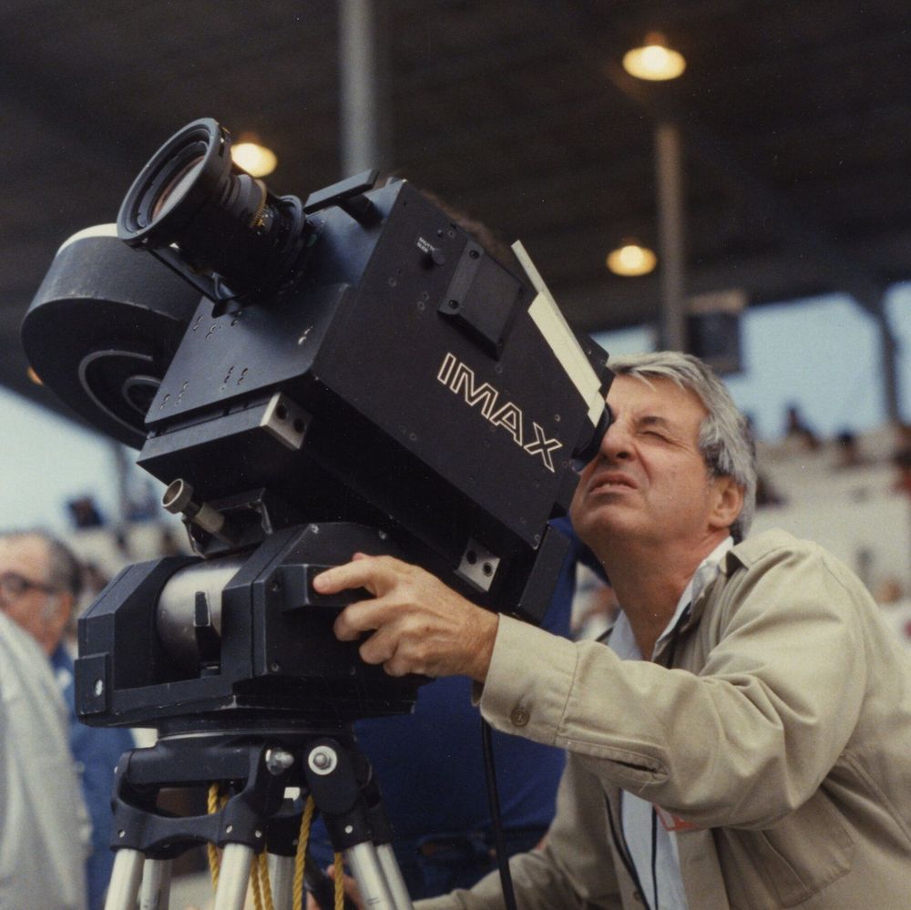 This black-and-white photograph captures IMAX co-founder and cinematographer Graeme Ferguson with gray hair intently peering through the viewfinder of a large IMAX camera mounted on a tripod, adjusting it with one hand. He wears a light-colored jacket and lanyard, set in an outdoor stadium with overhead lights, beams, and a blurred crowd in the background, evoking a 1980s film production scene.