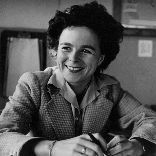 Black-and-white portrait of a smiling woman with short, curly dark hair, seated at a desk. She wears a checkered blazer over a blouse, a bracelet on her left wrist, and holds a pen in her right hand while resting her chin lightly on her left. Papers are scattered on the desk in front of her. The mood is warm and confident.