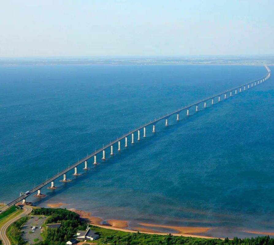 Aerial view of the Confederation Bridge, a long, curving two-lane bridge supported by numerous white concrete pillars, stretching across calm blue ocean waters from a green forested shoreline on the left toward a distant hazy landmass on the horizon under a clear light blue sky. The bridge appears to vanish into the distance over the sea.