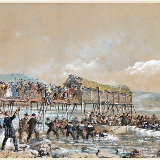 This is a 1866 oil painting by Robert Charles Dudley titled "Landing the Shore End of the Atlantic Cable." It depicts the arrival of the transatlantic telegraph cable at Heart's Content, Newfoundland, on July 27, 1866. A crowd in 19th-century attire cheers on a wooden pier over rocky shallows, with men wading in water to pull a rowboat ashore. A red flag waves from a scaffold on the left, and sod-roofed buildings stand on the pier amid a hilly, cloudy backdrop.