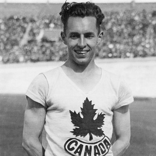 A young man with short, wavy hair smiles confidently at the camera. He wears a sleeveless athletic shirt with a large maple leaf and the word "CANADA" across the chest, along with a race bib numbered "667" pinned to his shorts. He stands on a track with a packed stadium of spectators in the blurred background. The black-and-white photo captures a moment from an early 20th-century Olympic or international athletic event.