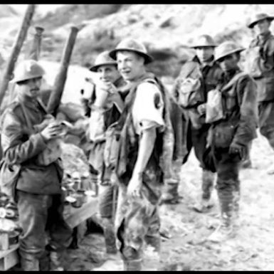 A black-and-white historical photo showing a group of soldiers in a rugged outdoor setting, likely a battlefield or trench area. Several soldiers are wearing helmets and military uniforms, with some operating or standing near artillery equipment. The ground is littered with debris, ammunition, and supplies, and the background features earthen fortifications or hills.