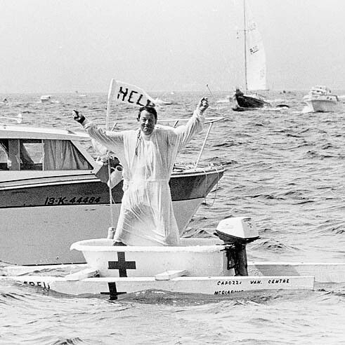A black-and-white photograph from the Nanaimo Bathtub Race depicts a smiling man dressed in a white robe standing in a motorized bathtub boat on open water. His arms are raised, holding a flag reading "HELP" in his right hand and a small pennant in his left. The bathtub features a large cross on the side, an outboard motor at the rear, and inscriptions "CARGILL VAN CENTRE" and "MICHIGAN." In the background, multiple boats—including a nearby motorboat marked "13K-44464" and distant sailboats—navigate the choppy sea.