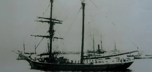 The image depicts an old sailing ship, likely from the late 19th or early 20th century, captured in a black-and-white photograph. The ship features multiple masts with rigging, characteristic of traditional sailing vessels of that era. The background shows a dock or harbor area, suggesting the ship is either moored or docked. The photograph's grainy texture and the ship's design indicate it might have been taken during a time when sail-powered ships were still common before the widespread use of steamships.
