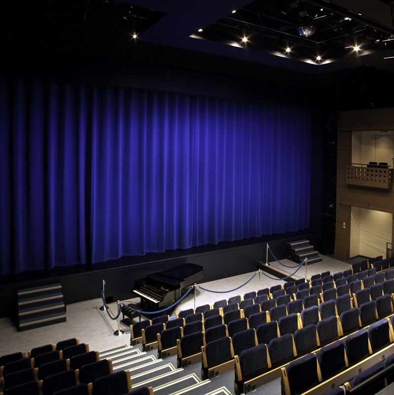 Interior photograph of an empty theater auditorium featuring a large blue stage curtain, a grand piano on the stage behind a velvet rope barrier, tiered rows of dark seats facing the stage, side balconies, and overhead spotlights.