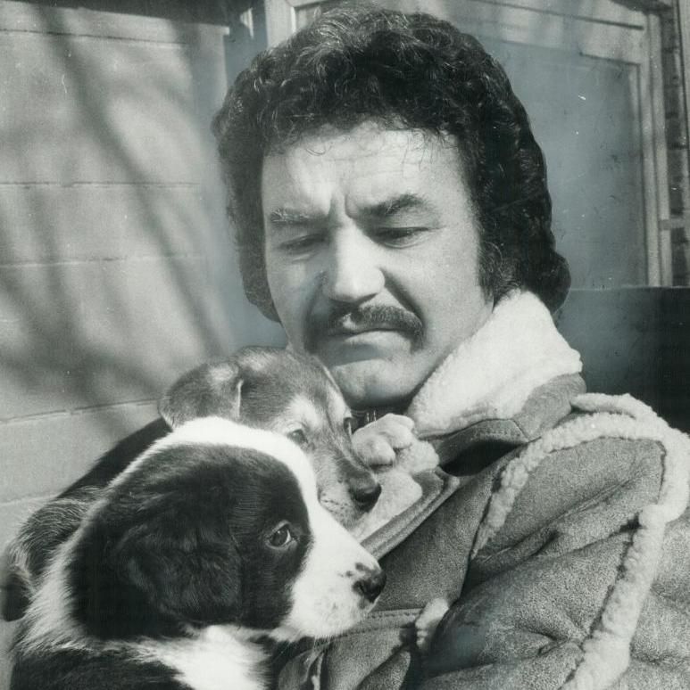 Black and white photo of a man with curly dark hair and a mustache, wearing a shearling jacket, holding a black-and-white border collie puppy close to his chest. The puppy looks at the camera while resting its head near the man's neck. They stand outdoors in front of a building with horizontal siding.