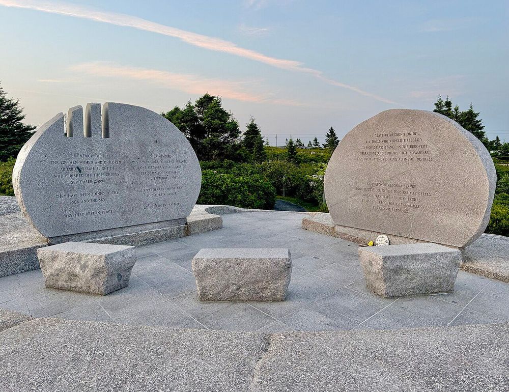 Two large stone monuments with inscriptions, set on a paved platform with three stone benches, overlooking a scenic landscape with trees and a clear sky at dawn or dusk. The left monument is dedicated "In Memory of the 229 men, women and children aboard Swissair Flight 111 who perished off these shores September 2, 1998," with additional text in English and French. The right monument offers a "Grateful recognition" to those who assisted in the recovery efforts, also in English and French. A small yellow flower and note are placed near the base of the right monument.