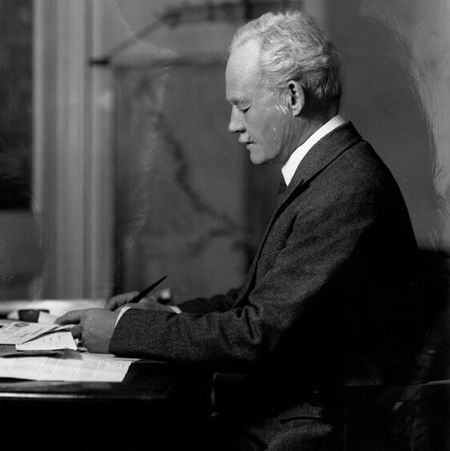 A black-and-white photograph of an older man with white hair, wearing a formal suit and tie, seated at a desk and writing or reviewing papers. The background includes a map or chart on the wall, and the setting appears to be an office or study with a professional atmosphere.