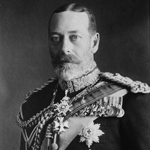 Black and white formal portrait of a man in ornate military dress uniform. He has short hair, a full beard, and wears epaulettes, a sash, multiple medals, stars, and chains across the chest. The background is plain dark gray. The image has a vintage tone with high detail in the decorations.