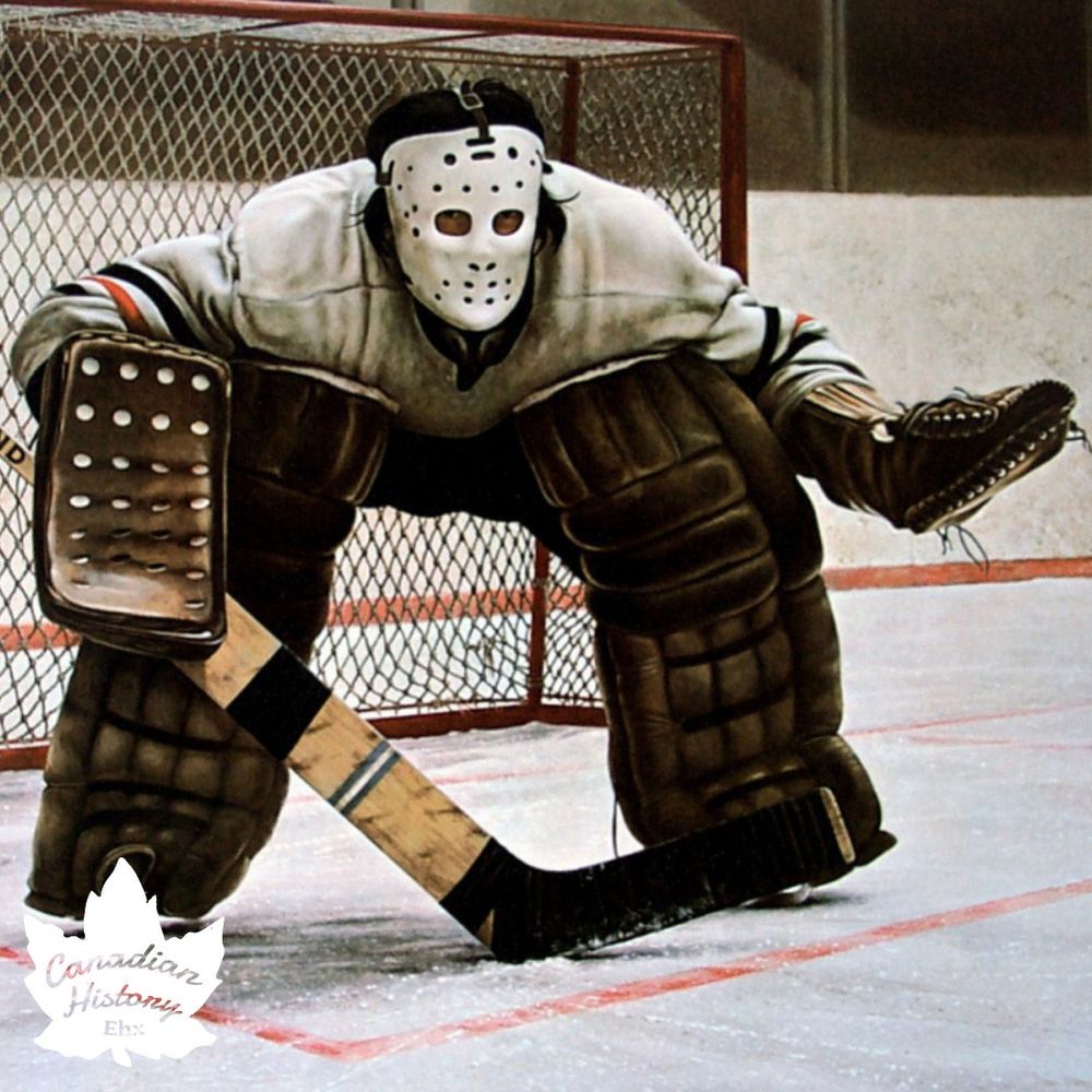 The photo depicts a hockey goalie in action, positioned in front of the net. The goalie is wearing classic, vintage-style equipment, including a white jersey with black and orange stripes, large brown leg pads, a catching glove, and a blocker. The mask is an older design, featuring a cage with multiple holes for visibility. The goalie stick is wooden with black tape on the blade. The setting is an ice hockey rink, with the goal net and ice surface visible. 