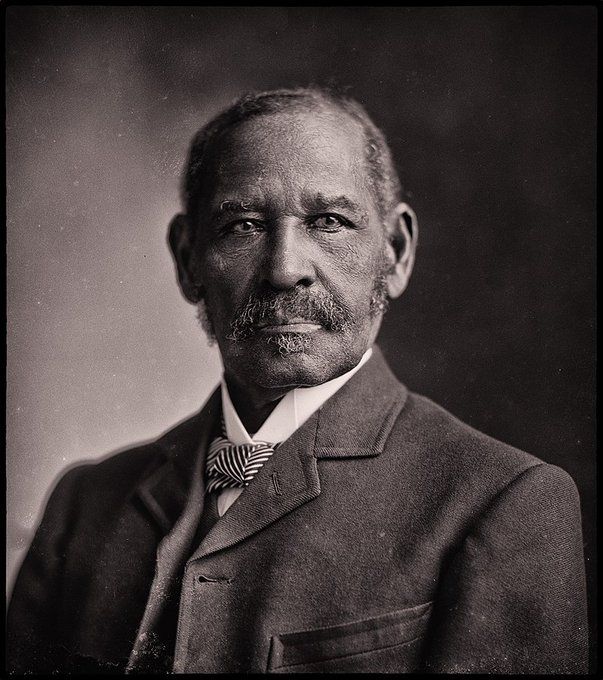 This is a black-and-white photograph of a man dressed in formal attire. He is wearing a suit jacket with a pocket on the left side, a white shirt, and a patterned bow tie. The photograph has a vintage feel, suggesting it might be from an earlier era, possibly the late 19th or early 20th century. The background is plain and dark, which helps to focus attention on the subject. 