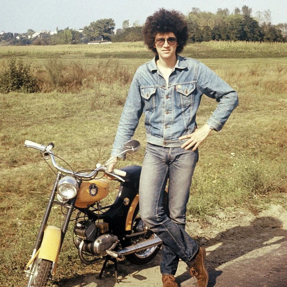 A young man with a large afro hairstyle and sunglasses, dressed in a denim jacket, jeans, and brown boots, posing with one hand on his hip and the other on a yellow and black motorcycle in a grassy rural field with trees and buildings in the background.