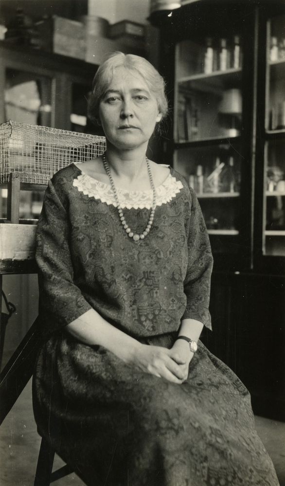 A black-and-white photo shows a woman in a patterned dress with a lace collar and pearl necklace, sitting in a chair. The background features shelves with bottles and a wire basket, suggesting an early 20th-century setting, possibly a pharmacy or study.
