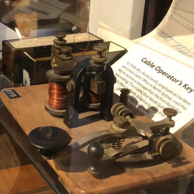 **Alt text:** Close-up of a museum display case showing a vintage telegraph key on a wooden base, labeled as a "Cable Operator's Key" used in 1918 by employee Annie Smith to receive the North Sydney message ending World War I. The key has brass knobs, coiled wires, and a lever. A descriptive card with text leans beside it. In the background, old documents and another electrical device are visible behind glass.
