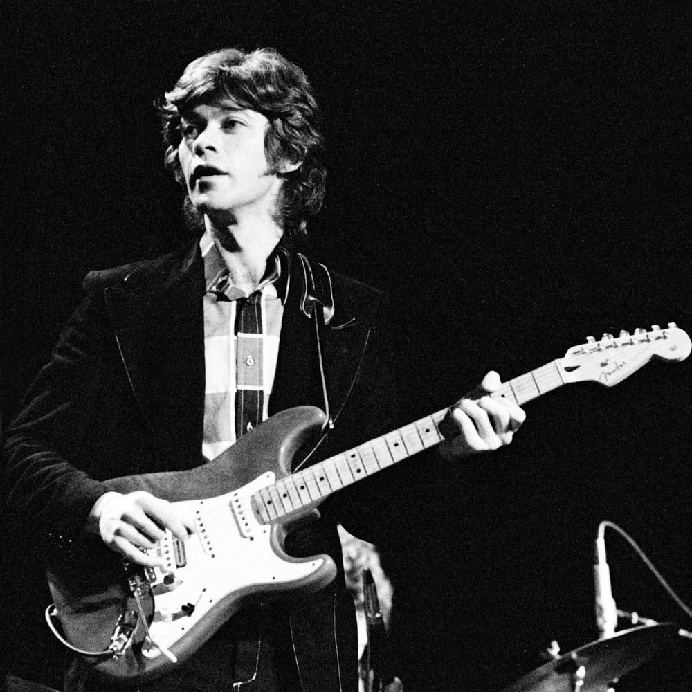 Black and white photograph of Robbie Robertson, a young man with shoulder-length wavy hair, performing on stage with his eyes closed and mouth open as if singing, playing a light-colored Fender Stratocaster electric guitar held at chest level, dressed in a dark velvet jacket over a striped collared shirt and tie, with a drum set partially visible in the background against a black backdrop.
