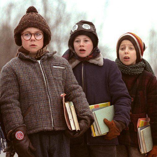 Three children stand outdoors in a snowy winter setting, bundled up in heavy coats, scarves, hats, and gloves. From left to right: the first child wears large round glasses, a brown pom-pom hat, plaid coat, and holds books; the second child wears goggles on their head, a dark coat with fur collar, and has an open-mouthed expression; the third child wears a striped knit hat, plaid coat, scarf, and holds stacked books while making a surprised face. Bare trees and snow are visible in the blurred background.