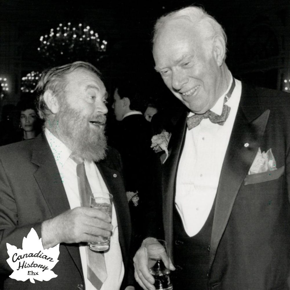 This black-and-white photograph captures a moment at what appears to be a formal event or gathering. Here's a detailed description:

People: The photo features two men engaged in conversation. The man on the left has a beard and is wearing a suit with a tie, holding a drink in his hand. The man on the right is dressed in a tuxedo with a bow tie and a boutonnière, also holding a drink. Both men are smiling, suggesting a friendly and light-hearted interaction.
Background: The background is dimly lit, typical of an indoor event setting, possibly a banquet or a gala. There are other attendees visible, dressed in formal attire, indicating the event's formal nature. A chandelier is visible in the background, adding to the elegance of the setting.