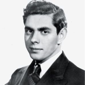 The photo is a black-and-white portrait of a young man with neatly styled, wavy hair. He is wearing a formal suit with a tie and a pocket square, presenting a polished and professional appearance. The background is plain, keeping the focus on his attire and composed expression.