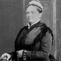 The black-and-white photo depicts a woman dressed in Victorian-era clothing, including a dark, elaborate dress with lace trim and a high collar. She wears a headpiece and a necklace with a pendant. The woman is standing with one hand resting on a cylindrical object, possibly a table or chair, against a plain backdrop.