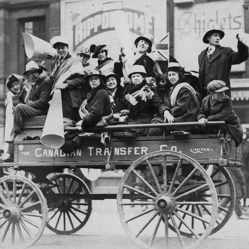 Black and white photograph of a group of people riding on top of a horse-drawn wagon labeled "THE CANADIAN TRANSFER CO. LIMITED." Men and women wear early 20th-century clothing including hats, coats, and scarves. They hold large paper megaphones and rolled banners, waving at the camera. A building in the background has signs reading "HIPPODROME" and "Tickets." The scene is on a city street with other pedestrians and vehicles visible. The image shows signs of age with scratches and grain.