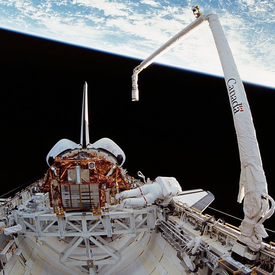 An astronaut in a spacesuit is working in space, positioned near the payload bay of a space shuttle with its cargo bay doors open. A large robotic arm labeled "Canada" extends from the shuttle, holding a payload. The Earth is visible in the background with a blue atmosphere and cloud formations against the blackness of space.
