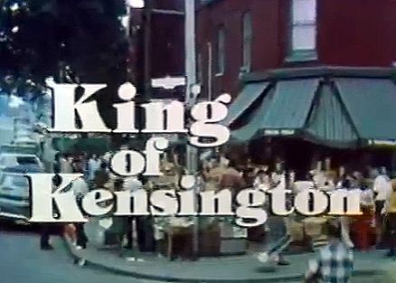 The photo appears to be a still from a television show or movie, featuring the title "King of Kensington" prominently displayed in large, bold white letters with a slight black outline. The background shows an outdoor street scene, likely set in an urban neighborhood. There are several people gathered around, some standing and others possibly interacting, with a mix of casual clothing styles suggesting a mid-20th-century setting, possibly the 1970s. The architecture includes a building with a green awning and a red brick facade, and there’s a streetlight and a vehicle visible, adding to the urban atmosphere. The overall tone of the image has a vintage, grainy quality, consistent with older film or TV productions.