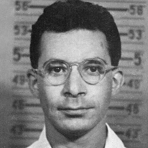 Black-and-white mugshot of a young man with short dark hair and round wire-framed glasses, facing forward with a neutral expression, standing against a height chart marked in inches from 4'6" to 5'9". He wears a light-colored collared shirt.