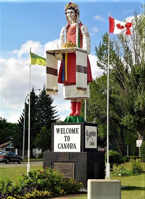 A large statue of a woman in traditional Ukrainian attire stands on a pedestal with a "Welcome to Canora" sign. She holds a loaf of bread and is surrounded by Canadian and Ukrainian flags, with greenery and a clear sky in the background.