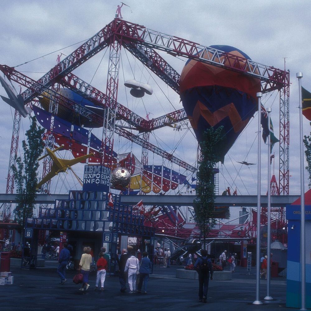 Color photo of a bustling fairground with a large hot air balloon and various aircraft suspended under a red and silver framework, featuring the "Expo '86" sign, with people walking and flags from different countries in the background under a cloudy sky.