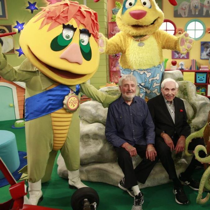 A colorful children's TV set with bright green walls, round windows, and toy decorations. Four characters pose together: a large yellow puppet with spiky orange hair, green eyes, and a blue sash stands on the left; a tall yellow dog puppet in blue bone-patterned shorts stands behind; two elderly men (one with gray hair and beard in a blue shirt, the other white-haired in a black suit) sit smiling on a fake rock in the center; a green octopus-like monster puppet with seaweed hair and tentacles sits on the right. Everyone appears cheerful in the playful room.