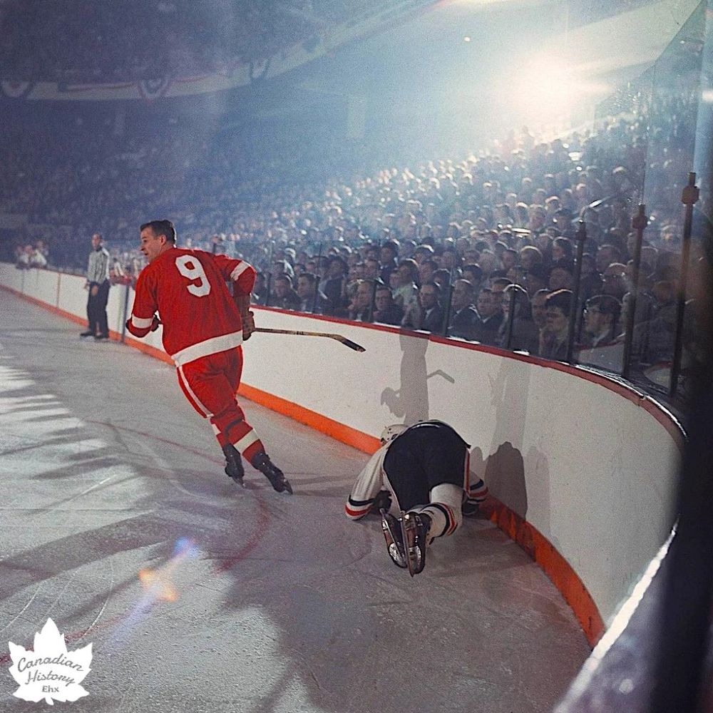 The photo depicts an intense moment during an ice hockey game, likely from a historical era given the style of the uniforms and the quality of the image. A player wearing a red jersey with the number 9 is skating on the ice, holding a hockey stick, and appears to be in motion, possibly pursuing the puck or maneuvering around an opponent. The red jersey features white stripes on the sleeves and pants, suggesting it could be from a team like the Detroit Red Wings.
Another player, wearing a dark jersey with orange and white accents, is on the ice