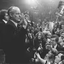 Black and white photo of a man with white hair, wearing a suit, speaking into multiple microphones at a crowded event, with a large audience holding signs and cheering. The scene is brightly lit with a lively atmosphere.