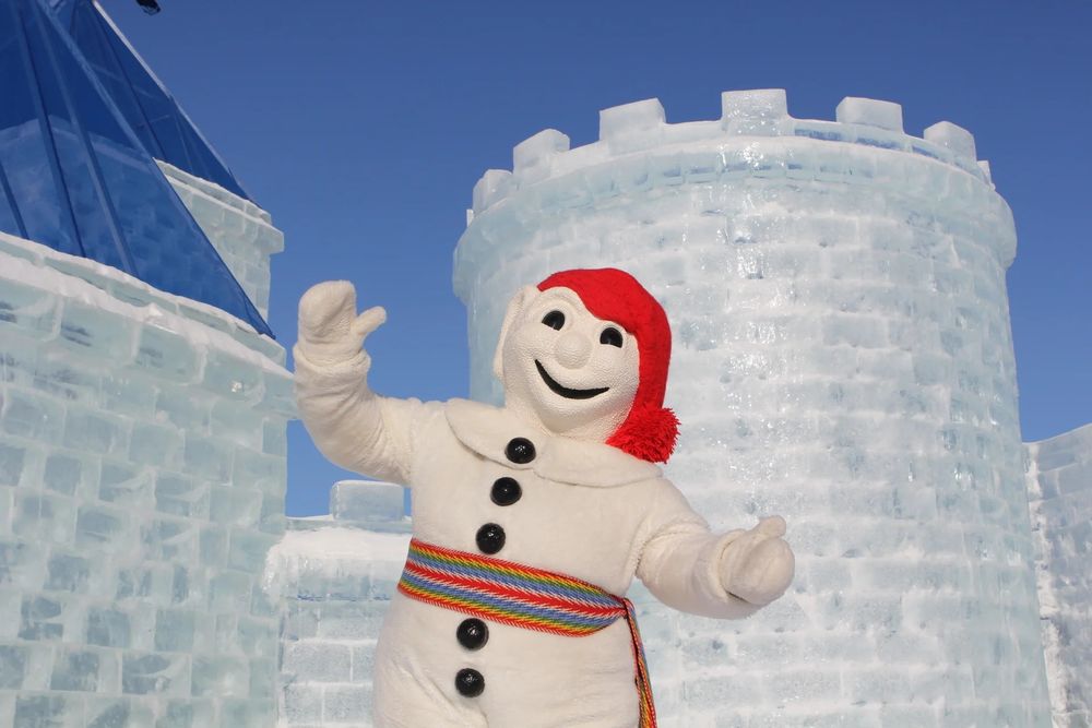 The photo features a person in a large, cheerful snowman costume standing in front of an impressive ice castle structure. The snowman costume is white with black button details and a colorful striped belt, and it's topped with a red hat with a tassel. The ice castle behind the snowman is intricately constructed from large blocks of ice, with a blue-tinted roof and cylindrical towers, giving it a majestic and fairy-tale-like appearance. The sky is clear and blue, suggesting a bright, sunny day. 