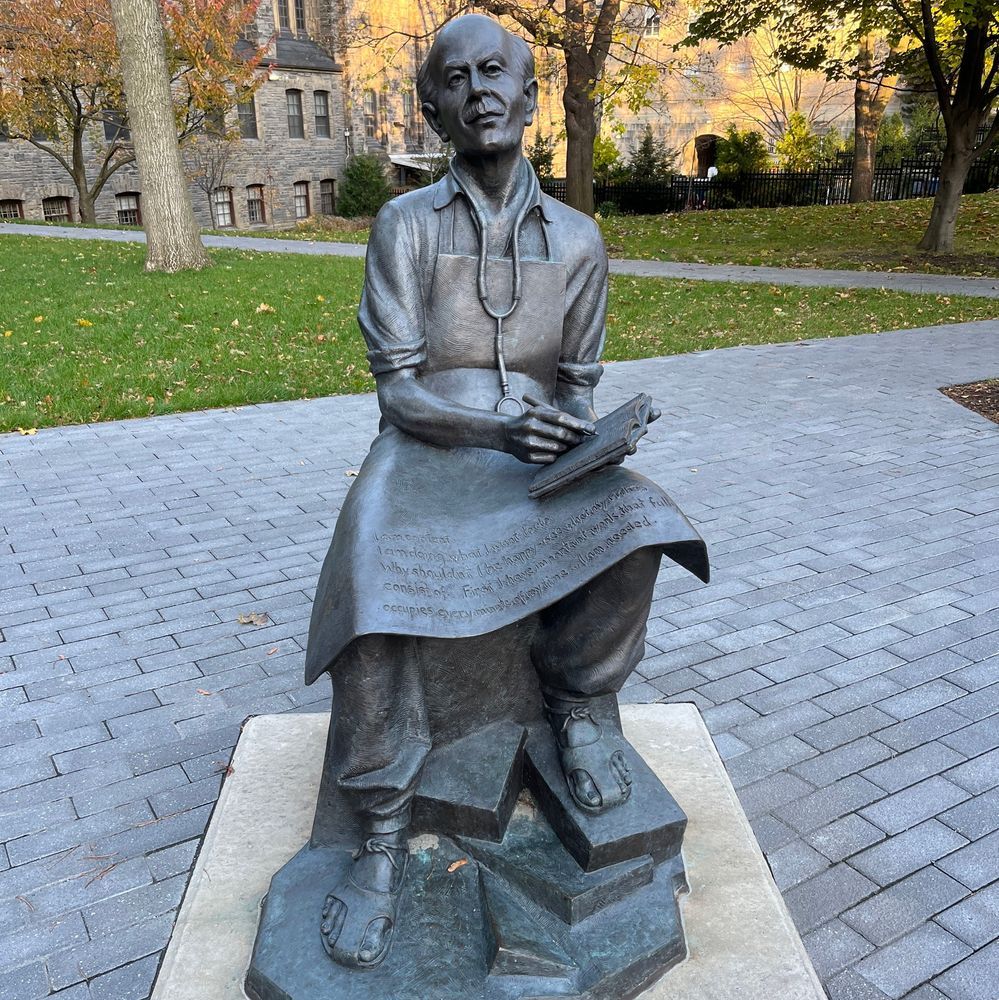 Color photo of a bronze statue of Norman Bethune on a stone pedestal in an outdoor park. He stands holding an open book in one hand and a scroll draped over his arm with inscribed text. Stethoscope around his neck, wearing a long coat. Background shows paved walkway, green grass, autumn trees, and a large stone building.