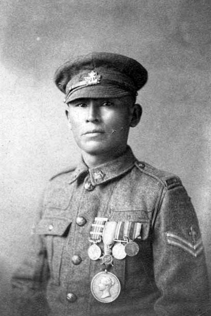 The photograph is a black-and-white portrait of a person dressed in a military uniform, likely from the early 20th century, possibly around the time of World War I. The individual is wearing a peaked cap adorned with a badge that appears to feature a maple leaf, suggesting a connection to Canadian forces. The uniform includes a buttoned jacket with a high collar and pockets, and the person has a sergeant's stripe on the sleeve, indicating a non-commissioned officer rank.
The chest of the uniform is decorated with several medals and ribbons, signifying military honors or campaigns. Prominently displayed is a large medal with a profile relief, possibly a Victoria Cross or another significant award, hanging from a ribbon. The background is plain and faded, typical of formal studio portraits from that era, which helps to focus attention on the subject's attire and decorations. The overall tone of the image is somber and dignified, reflecting the historical context of military service.