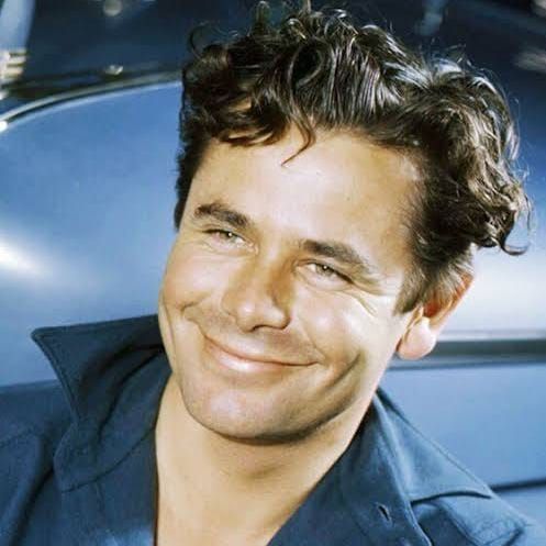 The photo shows Glenn Ford, a man with curly dark hair, wearing a blue collared shirt, and smiling slightly while leaning against a car.