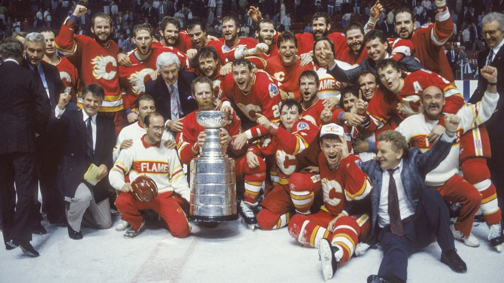 The photo shows a large group of hockey players from the Calgary Flames, identifiable by their red jerseys with a flaming "C" logo, posing on the ice with the Stanley Cup. They are celebrating, with some players holding the trophy, others raising their fists, and many smiling or cheering. The team is in full gear, including helmets and skates, and is surrounded by coaches and staff in suits. The background features a crowd of spectators in an arena, adding to the festive atmosphere. The image captures a moment of triumph, likely from their 1989 Stanley Cup victory.