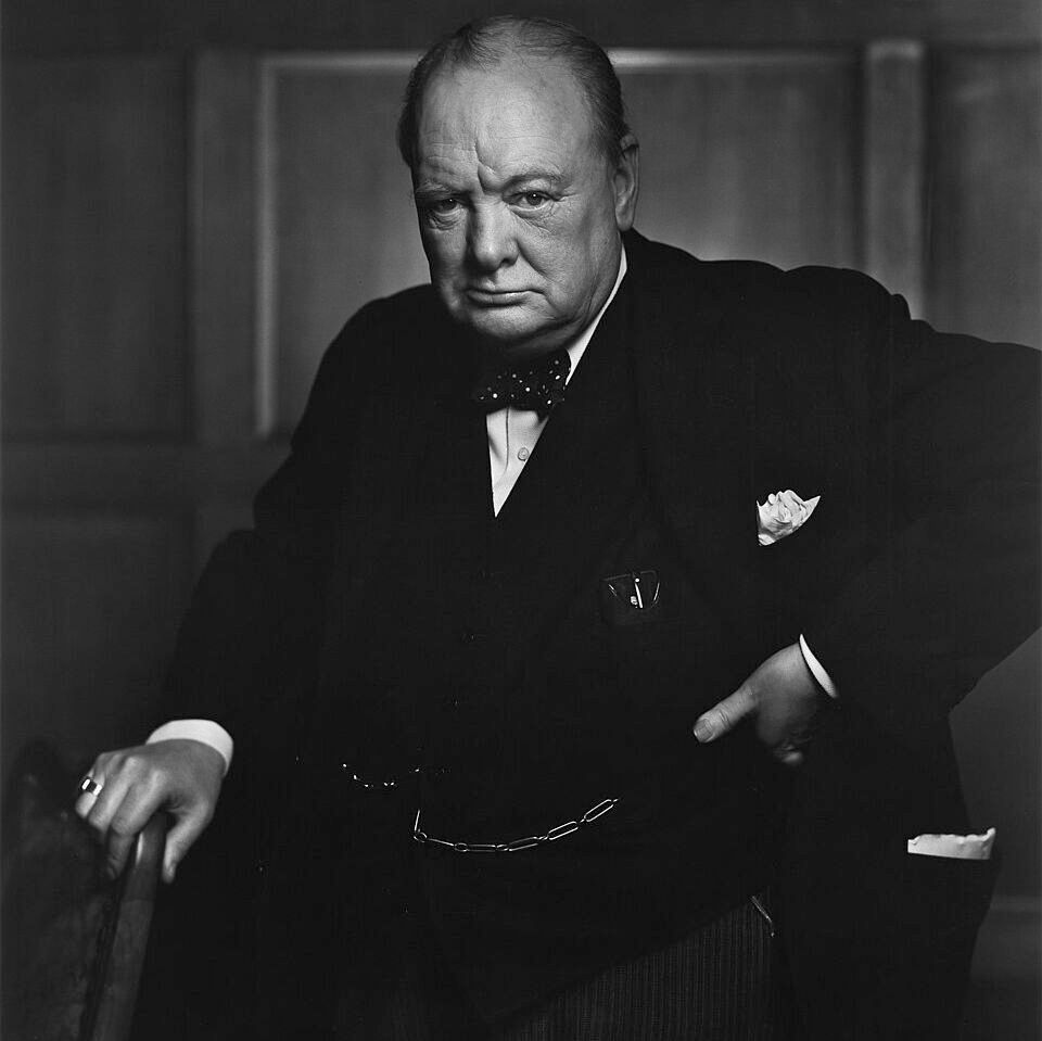 Black and white portrait photograph of Winston Churchill wearing a dark suit with bow tie, pocket square, and pocket watch chain. He has a stern expression, looking directly at the camera, with dramatic lighting casting shadows on his face and the paneled background. Taken by Yousuf Karsh in 1941.