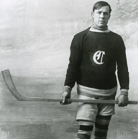 A black-and-white photo of a hockey player standing on skates, holding a hockey stick horizontally. He is wearing a dark sweater with a "C" logo, shorts, and striped knee-high socks with skates. The background is a faded, indistinct outdoor scene.