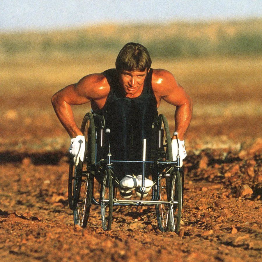 The photo shows Rick Hansen, a Canadian athlete and activist, propelling a wheelchair across a rugged, rocky terrain. He is wearing a black sleeveless top, white gloves, and athletic shoes, with a determined expression on his face. The background features a vast, open landscape under a clear sky.