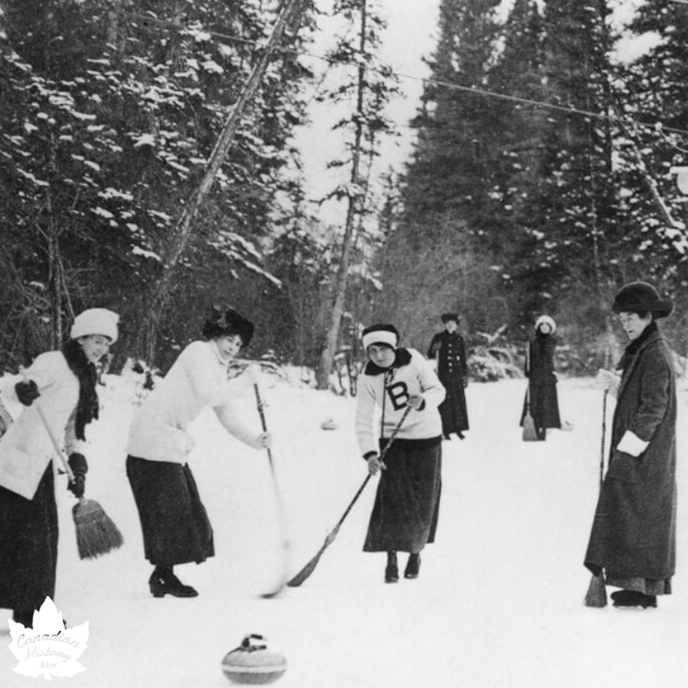 The photograph depicts a group of women playing curling on a snowy outdoor rink, surrounded by a forest of tall trees. The image is in black and white, suggesting it is historical. The women are dressed in early 20th-century winter attire, including long skirts, coats, hats, and scarves, with some wearing sweaters featuring a prominent letter "B." They are actively engaged in the game, using brooms to sweep the ice in front of a curling stone. The scene captures a moment of camaraderie and physical activity in a wintry, natural setting.