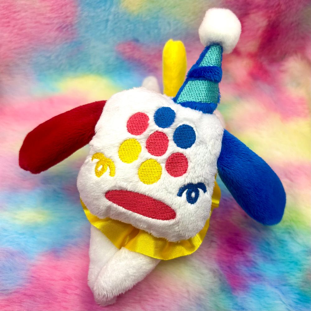 A white PUPP13 Plush with red and blue ears, blue red and yellow eyes, yellow and blue confetti cheeks, a red nose, and a yellow ruffle collar lays on a rainbow tie dye fur