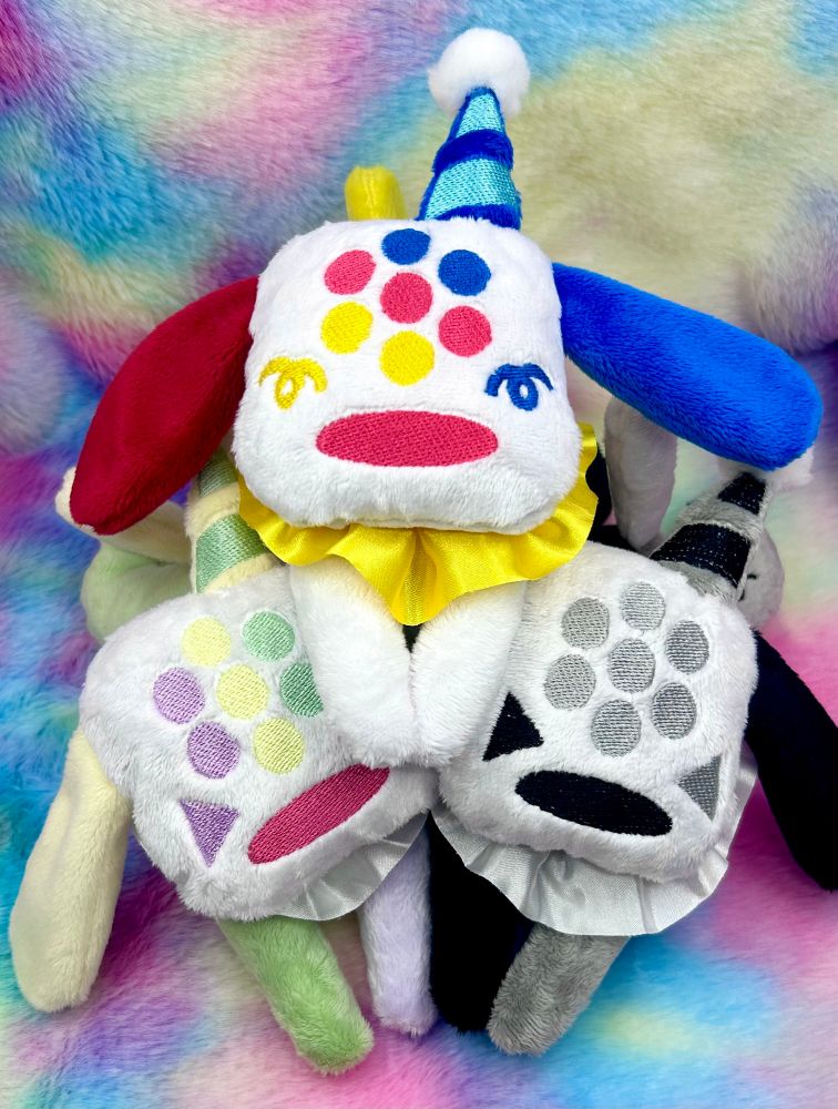 Three clown themed PUPP13 plushies. The one on top is white with yellow red and blue accents, the one on the bottom left is white with green purple and yellow accents and the one on the bottom right is white with black and gray accents